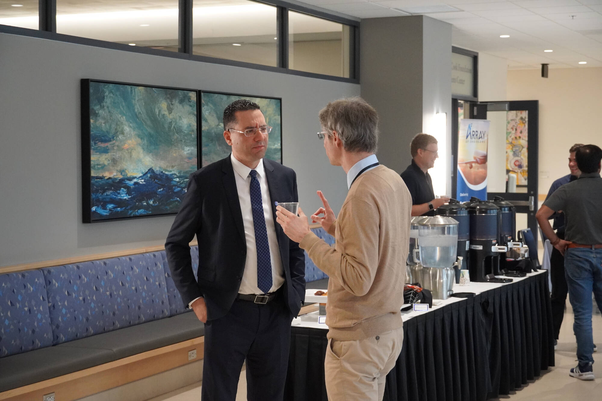 Dean, Marouane Kessentini and a speaker engage in conversation near a refreshment table during a networking break at an event.